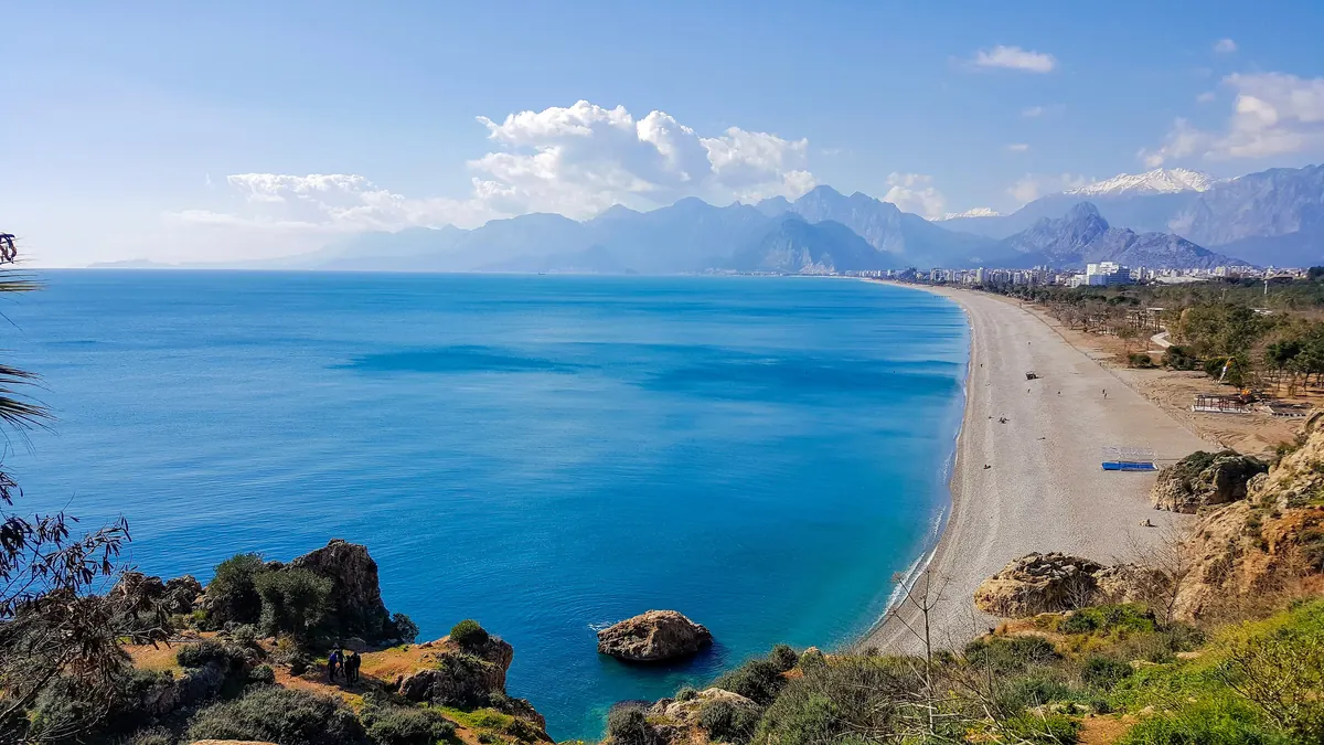 Plaja Konyaaltı din Antalya văzută de pe faleză: mare turcoaz, plajă lungă cu pietriș, orașul în depărtare și munții Bey cu vârfuri înzăpezite