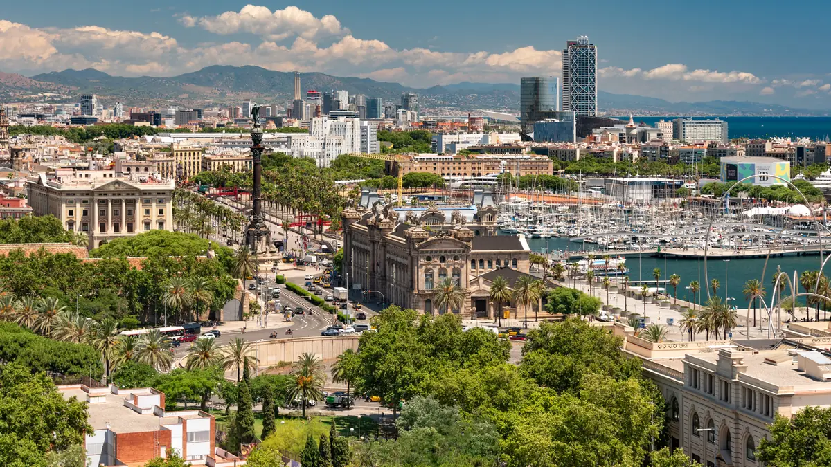 Barcelona văzută de sus: Monumentul lui Columb, portul Port Vell cu iahturi, clădiri vechi, palmieri și munții din fundal