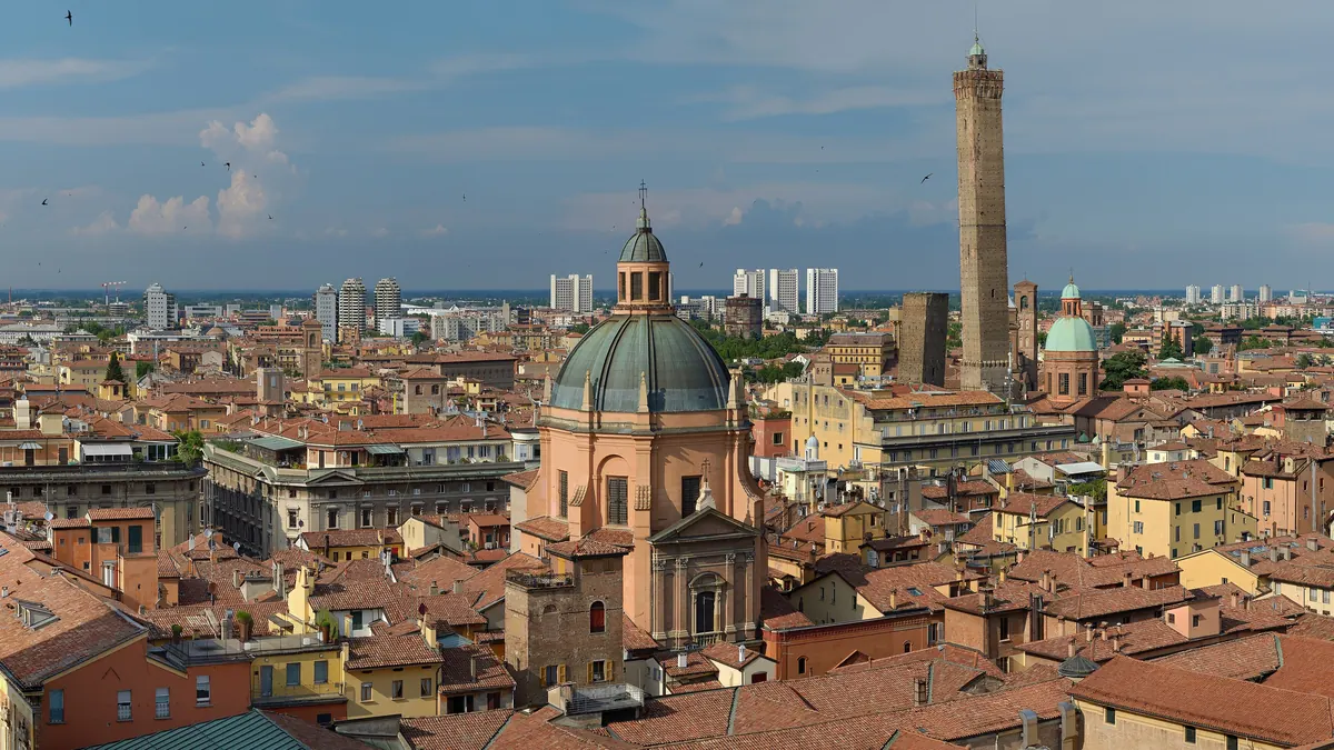 Acoperișurile roșii ale Bolognei văzute de sus, cu cupola sanctuarului Santa Maria della Vita în prim-plan și Turnul Asinelli în dreapta, sub un cer albastru cu nori