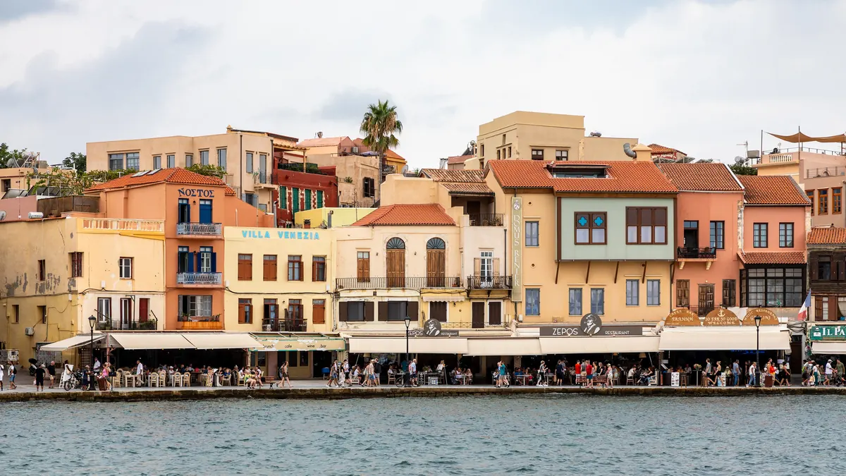 Portul venețian din Chania, Creta: case colorate cu acoperișuri de țiglă, terase de restaurant pe chei și apa portului în prim-plan
