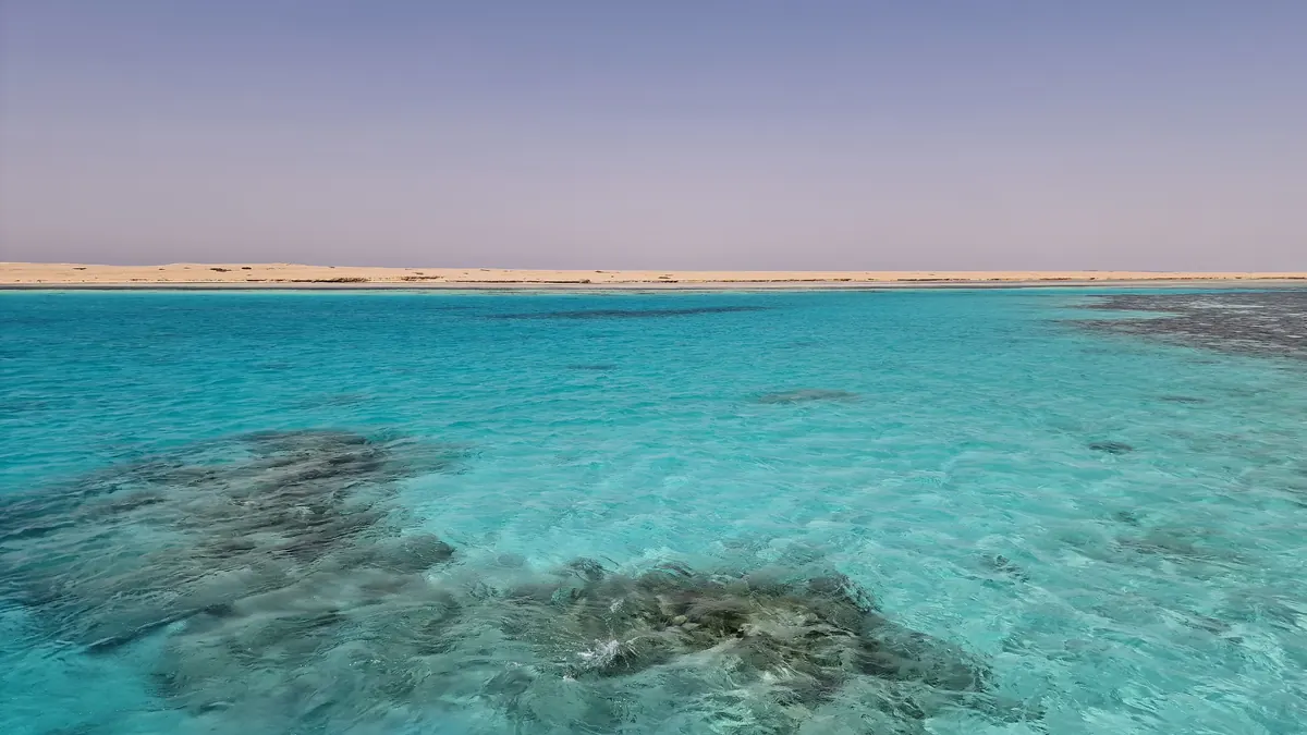 Apă turcoaz și recife de corali la mică adâncime în Marea Roșie, cu o insulă de nisip la orizont, lângă Hurghada