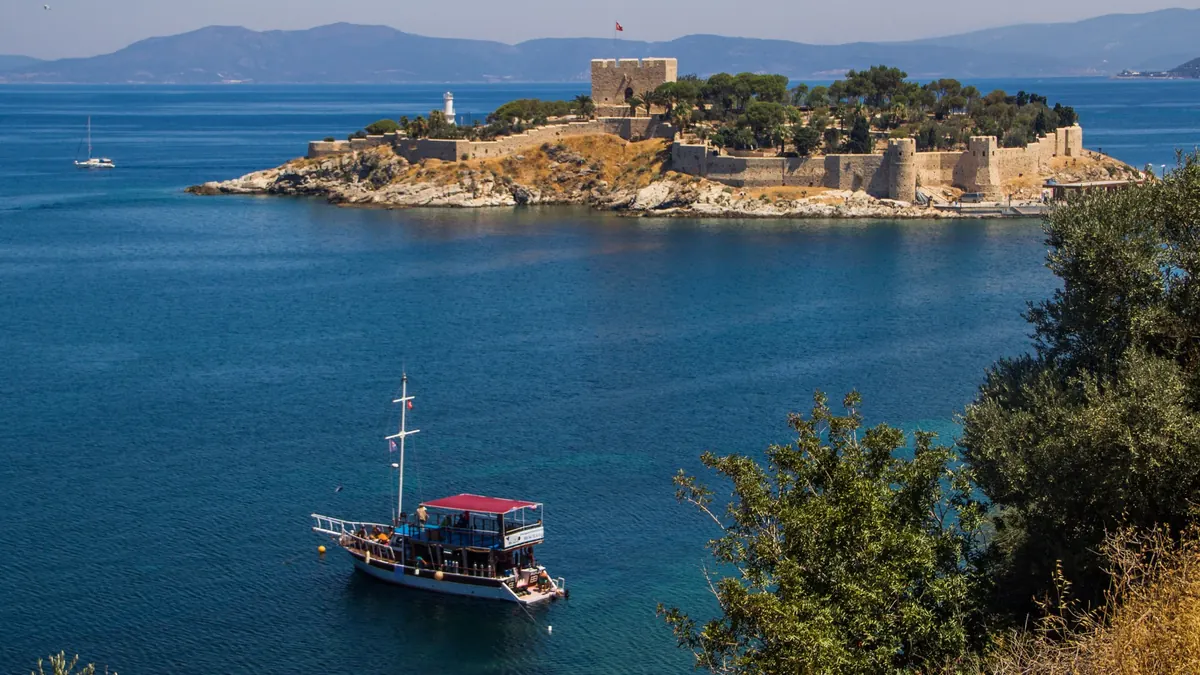 Insula Porumbeilor din Kușadası, cu cetatea de piatră și farul, marea albastră și un vas de excursie ancorat în golf
