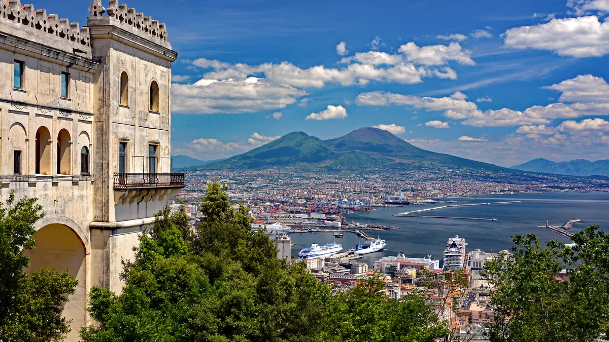 Golful Napoli și Vezuviul văzute de la Certosa di San Martino: clădirea mănăstirii, copaci, portul cu feriboturi și orașul la poalele vulcanului