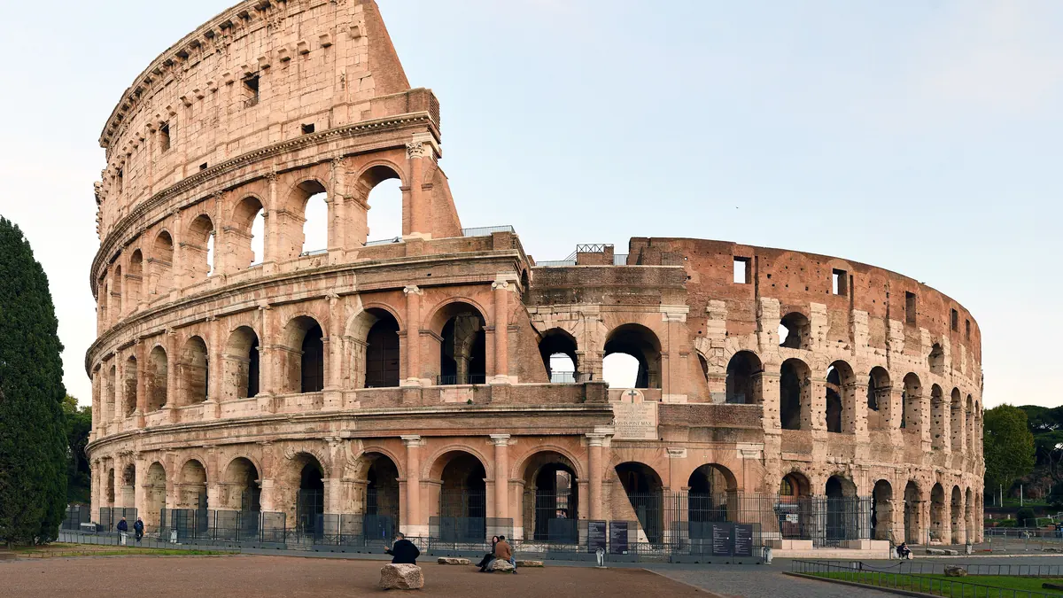 Colosseumul din Roma în lumina dimineții, cu arcadele din travertin, cerul senin și câțiva turiști în piața din fața lui