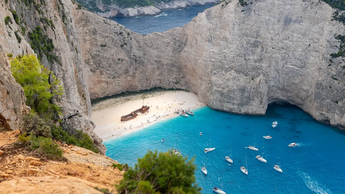 Plaja Navagio din Zakynthos văzută de pe faleză: epava Panagiotis pe nisipul alb, stânci calcaroase înalte și apă turcoaz cu bărci ancorate
